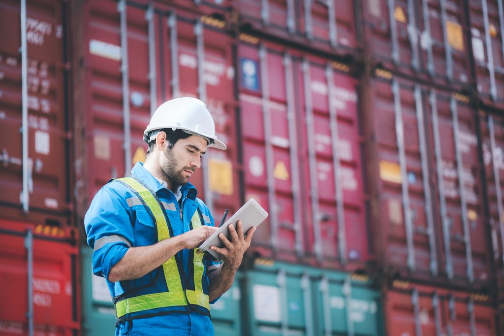 Company Security Officer wearing a high vis jacket and hard hat with a clipboard, stood in front of shipping containers.