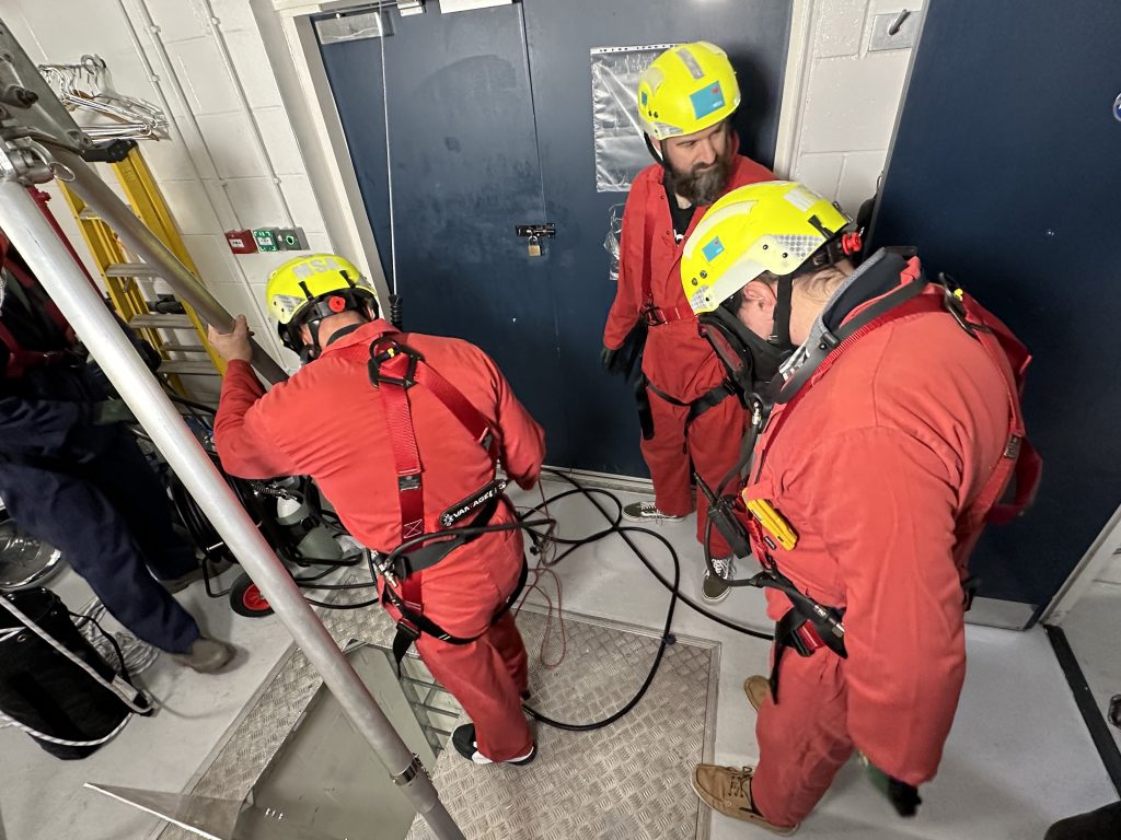 three delegates gathered around small trap door preparing for enclosed space entry