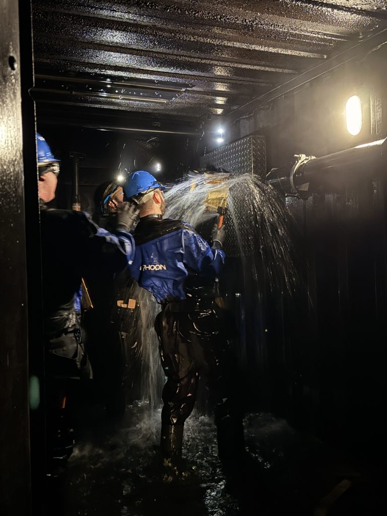 Maritime Damage Control Training at the Maritime Skills Academy in Kent. The image is of the inside of a container, posing as the interior of a ship's engine room that might experience a burst pipe or rupture. There are three crew members trying to stop the water flowing through a burst with wooden blocks. 